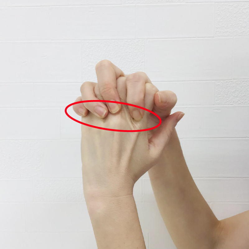 反対の手の指先が手の甲側の指の付け根（赤い丸の部分）に当たるように痛みのある側の指を握ります。